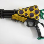 金と黒の銃型の品を側面から撮影。6つの装填部と緑のグリップがある。