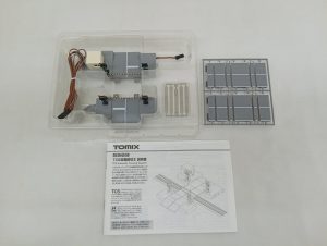 透明ケースに入った踏切のユニット2組と、灰色の部品の枠、TOMIXの説明書を並べた写真。